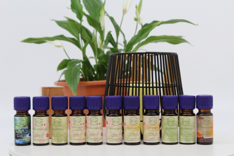 May include: A collection of 12 small glass bottles with purple lids, each containing essential oil. The bottles are arranged in a row on a white surface. The labels on the bottles feature a variety of designs and text in German.