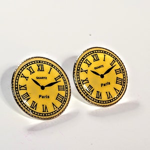 May include: A pair of gold-colored earrings with a clock face design. The clock face has black Roman numerals, black hands, and the words "QUARTZ" and "Paris".