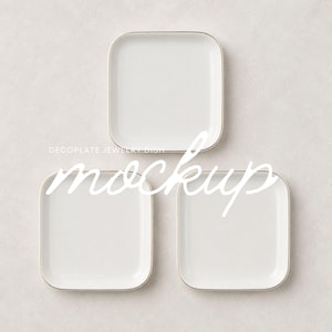 May include: Three square white jewelry dishes with rounded corners and thin metallic gold rims. The text DECOPLATE JEWELRY DISH MOCKUP is visible across the center of the three dishes, which are arranged in a triangular formation on a light surface.