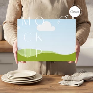 May include: A rectangular canvas mockup with a blue sky and green landscape design, with the word "MOCK UP" in white. The canvas is held by a person. A stack of plates and a folded napkin are on a wooden surface.