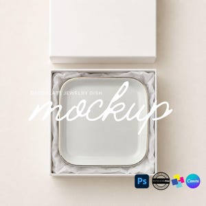 May include: A square white jewelry dish with a thin metallic rim, nestled inside a white gift box lined with white tissue paper. The text DECOPLATE JEWELRY DISH mockup is overlaid in white script across the center of the dish.