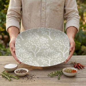 May include: An oval serving platter with a light green background and a white tree branch pattern. The platter is being held by a person. The image also shows small bowls of spices and herbs on a wooden table.