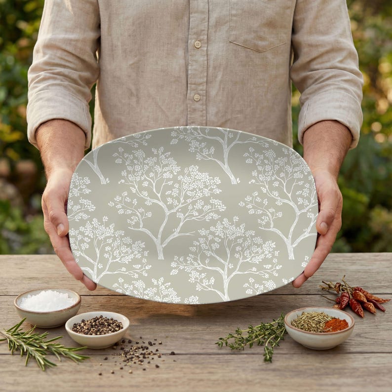 May include: An oval serving platter with a light green background and a white tree branch pattern. The platter is being held by a person. The image also shows small bowls of spices and herbs on a wooden table.