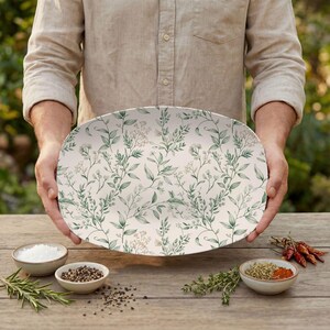 May include: An oval serving platter with a botanical design. The platter features a cream-colored background with a repeating pattern of green leaves and delicate white flowers. The platter is held over a wooden table with small bowls of spices.