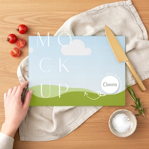 May include: A rectangular cutting board with a blue sky and green grass design, the word "MOCK UP" is printed on the board. A gold knife, tomatoes, rosemary, and a small bowl of salt are arranged on a wooden surface.