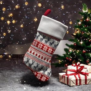 Op de afbeelding: Een rode en witte kerstsok met een grijs en wit sneeuwvlokkenpatroon en rendierontwerpen. De sok hangt aan een schoorsteenmantel met een kerstboom op de achtergrond.