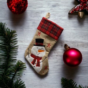 May include: A beige Christmas stocking with a snowman design. The stocking has a red and black plaid cuff and is decorated with white snowflakes.