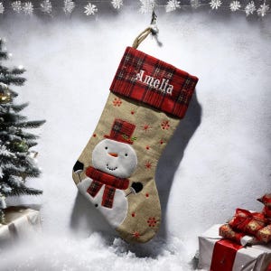 May include: A personalized Christmas stocking with a snowman design. The stocking is made of burlap and has a red plaid cuff with the name "Amelia" embroidered on it.