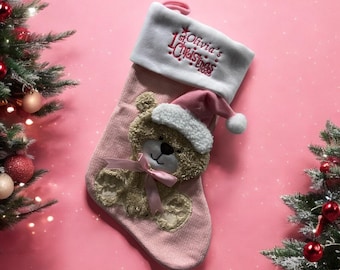 May include: Pink knitted Christmas stocking with a plush teddy bear wearing a Santa hat. The stocking has white felt trim with the text "1st Olivia's Christmas 2023" embroidered in pink.