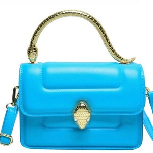 May include: A bright blue handbag featuring a structured rectangular body with a smooth finish. The bag includes a gold-toned metal handle shaped like a snake and a matching snake-head clasp on the front flap. It also has a detachable shoulder strap.