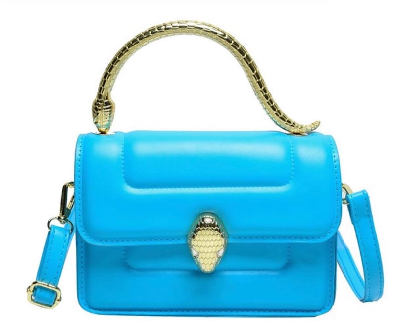 May include: A bright blue handbag featuring a structured rectangular body with a smooth finish. The bag includes a gold-toned metal handle shaped like a snake and a matching snake-head clasp on the front flap. It also has a detachable shoulder strap.