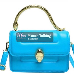 May include: A bright blue handbag featuring a gold-toned snake-shaped handle and a matching snake head clasp on the front flap. The bag includes a detachable shoulder strap and displays the text Mknoe Clothing mknoe.com on the front surface.