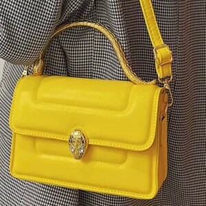 K&ouml;nnte beinhalten: Eine leuchtend gelbe, rechteckige Handtasche mit gesteppter Textur, einem goldfarbenen Schlangenkopf-Verschluss mit eingesetzten Kristallen, einem geschwungenen Tragegriff mit strukturierter Oberfl&auml;che und einem verstellbaren Schulterriemen.