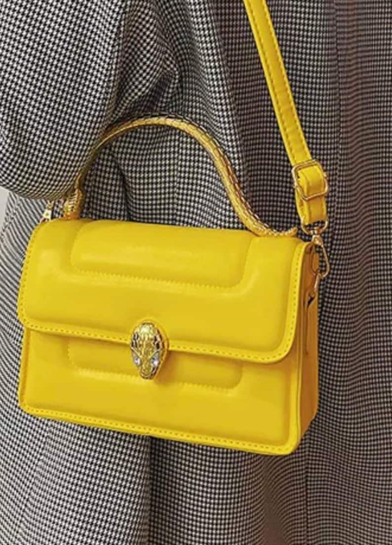 K&ouml;nnte beinhalten: Eine leuchtend gelbe, rechteckige Handtasche mit gesteppter Textur, einem goldfarbenen Schlangenkopf-Verschluss mit eingesetzten Kristallen, einem geschwungenen Tragegriff mit strukturierter Oberfl&auml;che und einem verstellbaren Schulterriemen.