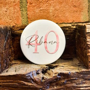 Könnte beinhalten: Ein weißer runder Knopf mit der Zahl 40 in Rosa und dem Namen Ribana in Kursivschrift.
