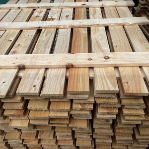 May include: A stack of unfinished wooden pallets. The pallets are made of light-colored wood with visible grain and knots. The boards are arranged in a grid pattern, with horizontal and vertical planks.