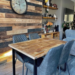 May include: A rustic wooden dining table with a black metal frame and four black upholstered chairs. The table has a distressed wood finish and the chairs have a quilted design. The table is set against a wall with reclaimed wood panelling.
