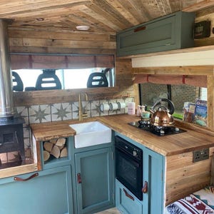 May include: A small kitchen inside a converted van with teal green cabinets, a wood countertop, a white porcelain sink, a black oven, and a wood-burning stove. The walls and ceiling are covered in reclaimed wood.