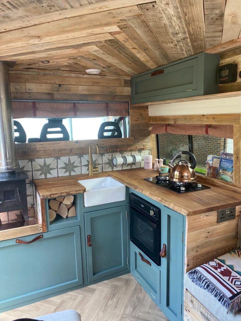 May include: A small kitchen inside a converted van with teal green cabinets, a wood countertop, a white porcelain sink, a black oven, and a wood-burning stove. The walls and ceiling are covered in reclaimed wood.