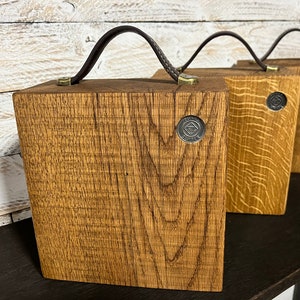 Puede incluir: Tres cajas cuadradas de madera con vetas visibles, cada una con un asa de cuero marr&oacute;n oscuro sujeta con herrajes de tono lat&oacute;n. Un peque&ntilde;o emblema met&aacute;lico circular con el texto "FUNK LUMBER" est&aacute; incrustado en el frente de cada caja.