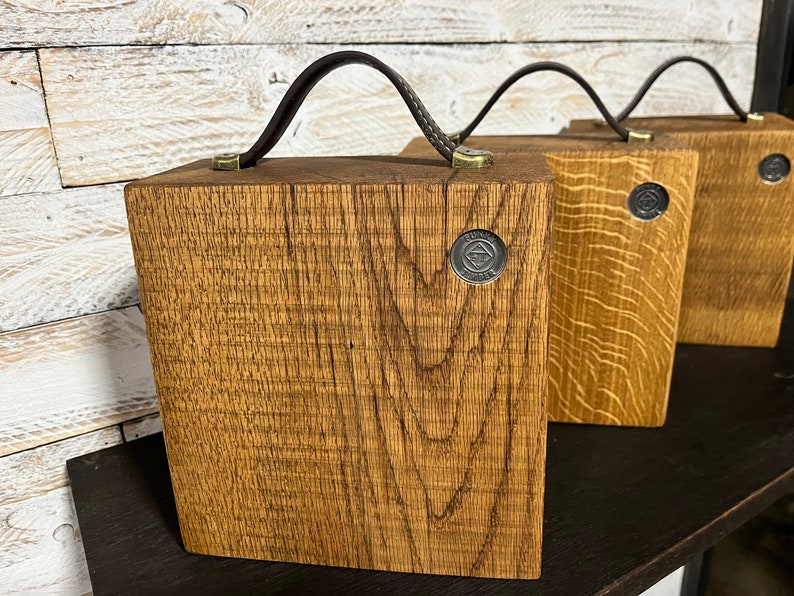 Puede incluir: Tres cajas cuadradas de madera con vetas visibles, cada una con un asa de cuero marr&oacute;n oscuro sujeta con herrajes de tono lat&oacute;n. Un peque&ntilde;o emblema met&aacute;lico circular con el texto "FUNK LUMBER" est&aacute; incrustado en el frente de cada caja.