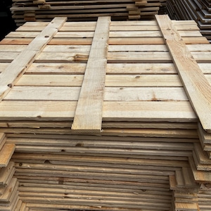 May include: A stack of unfinished wooden pallets, constructed from light-colored planks. The pallets are rectangular, with horizontal boards and vertical supports. The wood grain is visible, and the pallets appear ready for use in shipping or construction.