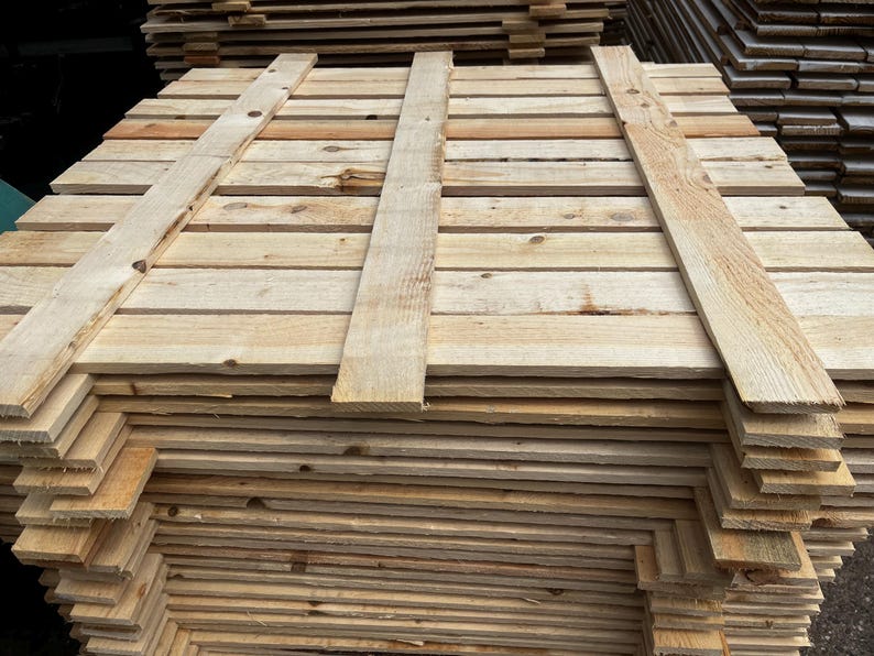 May include: A stack of unfinished wooden pallets, constructed from light-colored planks. The pallets are rectangular, with horizontal boards and vertical supports. The wood grain is visible, and the pallets appear ready for use in shipping or construction.
