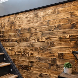 May include: A wall covered in horizontal, stained wooden planks with visible grain patterns and dark charred edges. To the left, a staircase with dark grey metal sides and wooden treads. A small potted plant sits on a surface in the foreground.