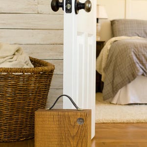 Puede incluir: Un tope de puerta cuadrado de madera con un asa de cuero oscuro y un detalle met&aacute;lico circular en la parte frontal. El tope est&aacute; colocado sobre un suelo de madera cerca de una puerta blanca con un pomo de metal oscuro y una cesta de mimbre.