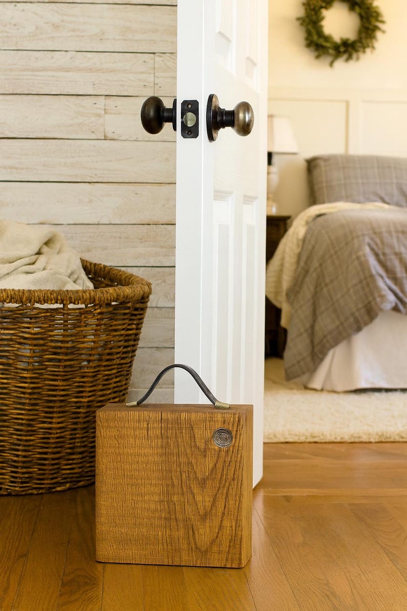 Puede incluir: Un tope de puerta cuadrado de madera con un asa de cuero oscuro y un detalle met&aacute;lico circular en la parte frontal. El tope est&aacute; colocado sobre un suelo de madera cerca de una puerta blanca con un pomo de metal oscuro y una cesta de mimbre.