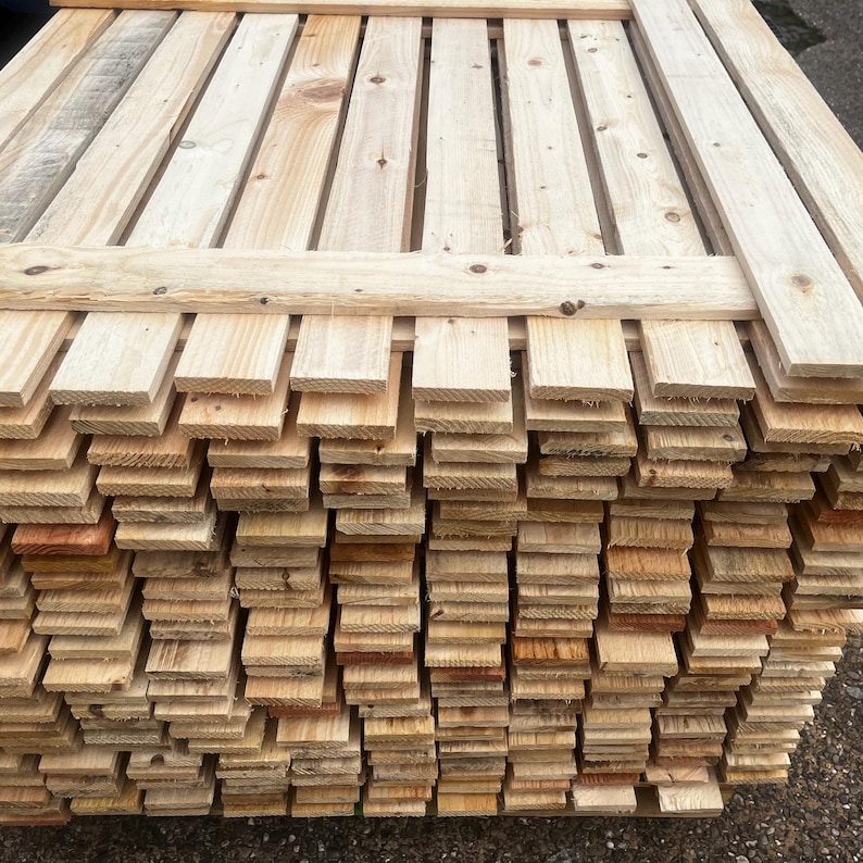 May include: A stack of unfinished wooden planks, varying in shades of light brown and tan. The planks are arranged in a neat, organized pile, with some planks forming a top layer. The wood appears to be new and ready for use in a construction or craft project.