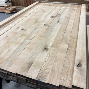 May include: A stack of light brown wooden planks, probably oak, with a natural wood grain pattern. The planks are arranged in a neat pile, ready for woodworking projects.
