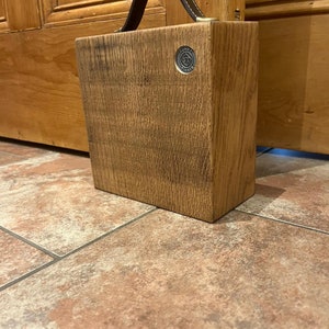 Puede incluir: Un bloque de madera rectangular con acabado de veta natural, que cuenta con un asa de cuero marr&oacute;n oscuro sujeta en la parte superior. Un peque&ntilde;o emblema met&aacute;lico circular est&aacute; incrustado en la esquina superior derecha con el texto Funky Timber.
