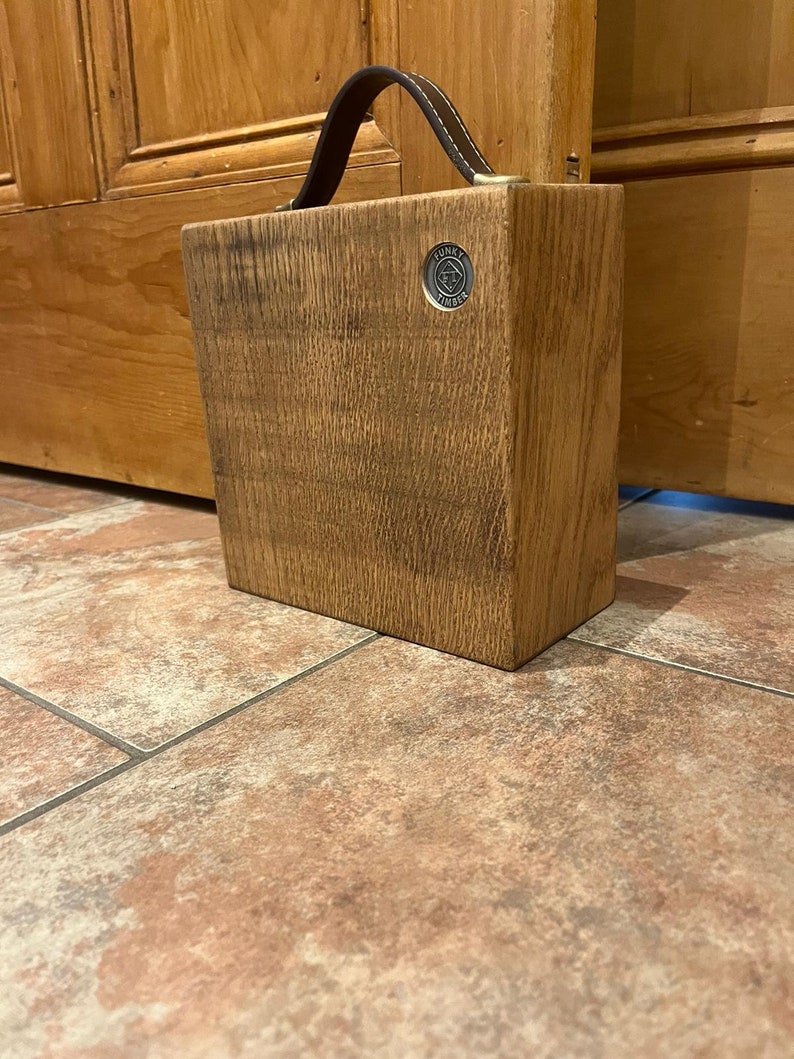 Puede incluir: Un bloque de madera rectangular con acabado de veta natural, que cuenta con un asa de cuero marr&oacute;n oscuro sujeta en la parte superior. Un peque&ntilde;o emblema met&aacute;lico circular est&aacute; incrustado en la esquina superior derecha con el texto Funky Timber.