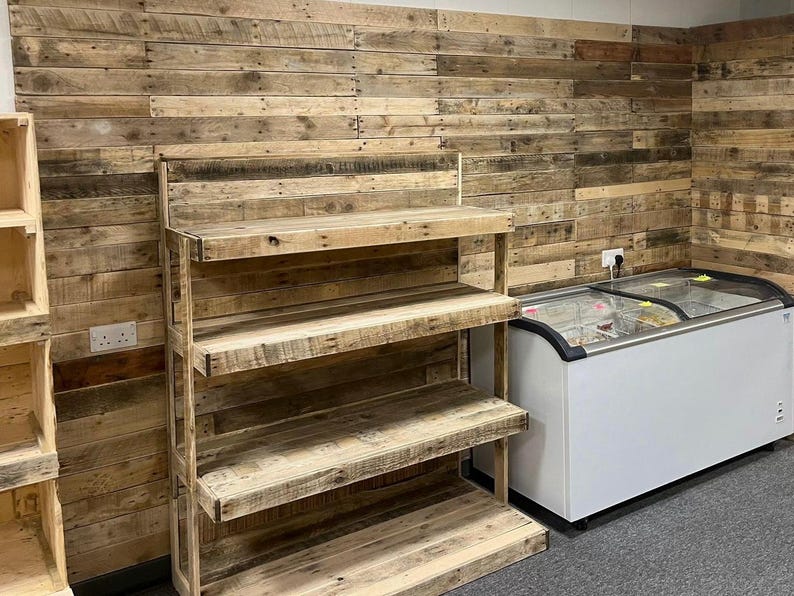 May include: A wooden shelf unit with three shelves, made from reclaimed wood planks, is set against a wall covered in a similar wood paneling. A white freezer with a glass top is in the background.