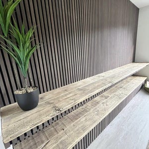 May include: A wall feature composed of dark brown vertical wooden slats with two integrated, light-toned rustic wooden shelves extending horizontally. A small black pot containing a green artificial plant sits on the top shelf surface.