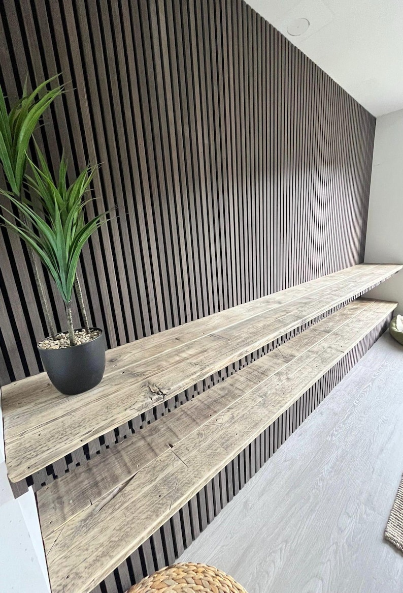 May include: A wall feature composed of dark brown vertical wooden slats with two integrated, light-toned rustic wooden shelves extending horizontally. A small black pot containing a green artificial plant sits on the top shelf surface.