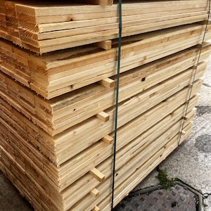 May include: A stack of unfinished wooden planks, tightly bound with a green strap. The planks are a light, natural wood color, showing the grain and texture of the material. The stack is neatly arranged, ready for construction or woodworking projects.