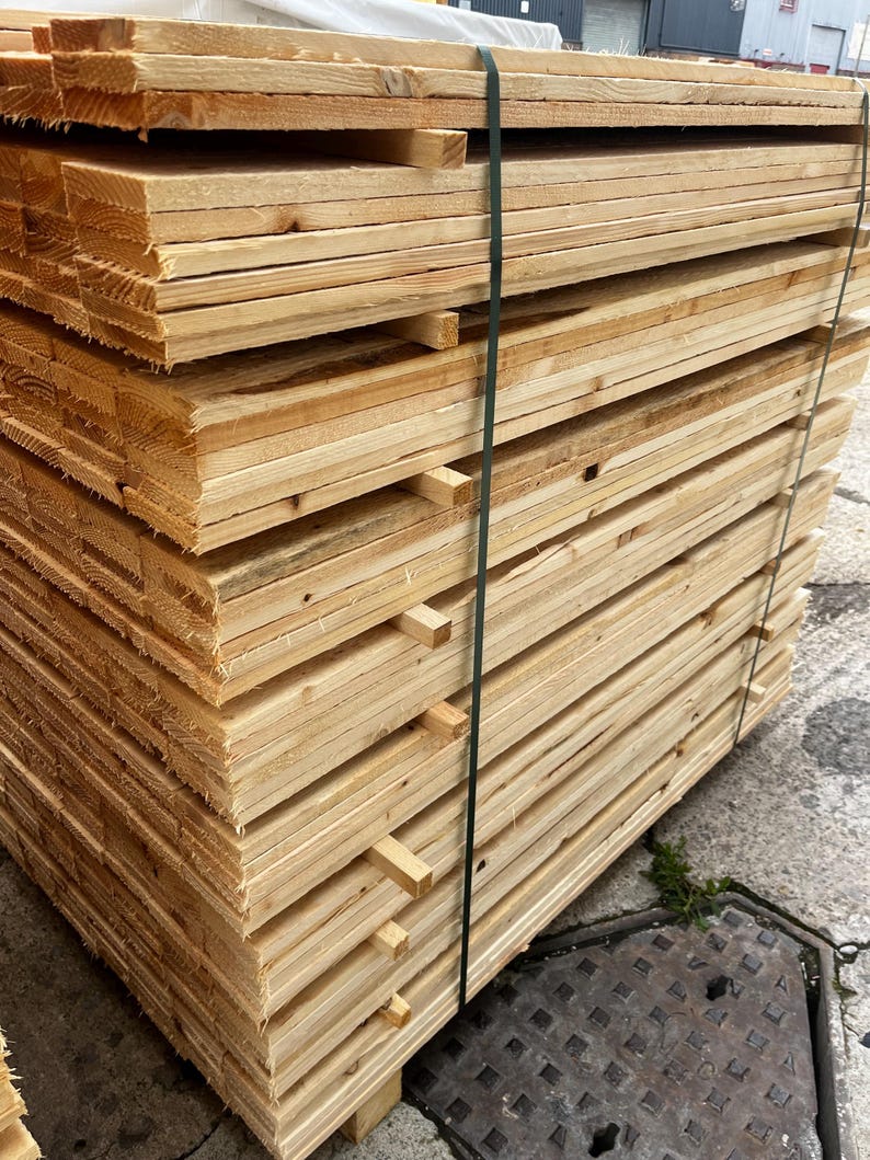 May include: A stack of unfinished wooden planks, tightly bound with a green strap. The planks are a light, natural wood color, showing the grain and texture of the material. The stack is neatly arranged, ready for construction or woodworking projects.