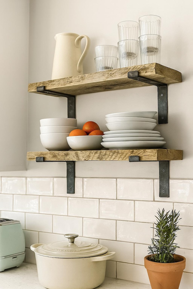 May include: Two natural wood shelves with dark metal brackets mounted on a wall. The top shelf holds a cream pitcher and clear ribbed glasses. The bottom shelf holds white bowls, orange citrus fruits, and a stack of white plates.