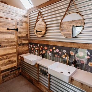 May include: A bathroom with rustic wood paneling and corrugated metal walls. Two round mirrors hang above a double sink with white porcelain basins. A floral wallpaper design is behind the sinks.