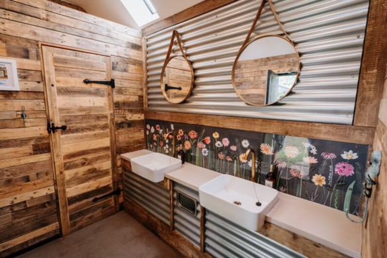 May include: A bathroom with rustic wood paneling and corrugated metal walls. Two round mirrors hang above a double sink with white porcelain basins. A floral wallpaper design is behind the sinks.