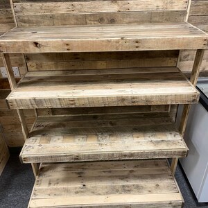 May include: A four-tier wooden shelf unit made from reclaimed wood planks. The shelves are stained in a natural wood color and have a rustic look. The shelf unit is freestanding and has a simple design.