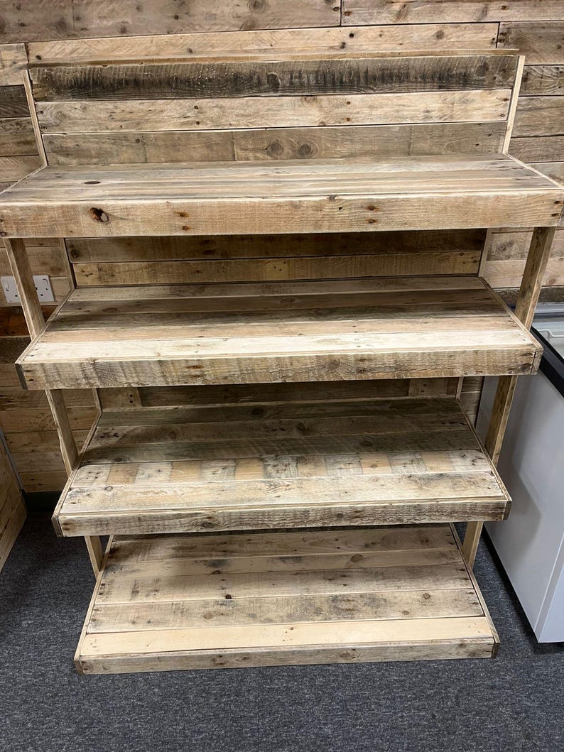 May include: A four-tier wooden shelf unit made from reclaimed wood planks. The shelves are stained in a natural wood color and have a rustic look. The shelf unit is freestanding and has a simple design.