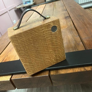 Puede incluir: Un bloque de madera cuadrado con veta visible, que cuenta con un asa de cuero marr&oacute;n oscuro sujeta con herrajes de color lat&oacute;n. Un peque&ntilde;o emblema met&aacute;lico circular est&aacute; incrustado en la esquina superior derecha con el texto Funky Timber.
