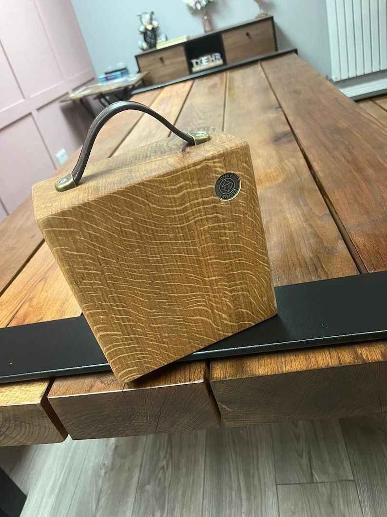 Puede incluir: Un bloque de madera cuadrado con veta visible, que cuenta con un asa de cuero marr&oacute;n oscuro sujeta con herrajes de color lat&oacute;n. Un peque&ntilde;o emblema met&aacute;lico circular est&aacute; incrustado en la esquina superior derecha con el texto Funky Timber.