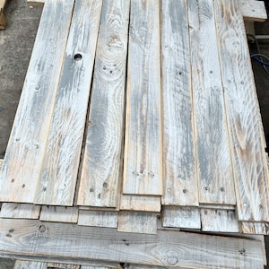 May include: A stack of weathered wood planks, painted white with grey accents. The planks are arranged in a staggered pattern, creating a rustic and distressed look.