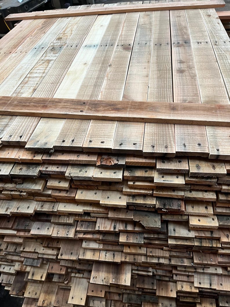 May include: A stack of reclaimed wood planks, some of which are nailed together to form a larger panel. The wood is a mix of light and dark brown tones, with a weathered and rustic appearance.