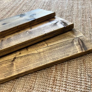 May include: Three rectangular wooden boards with a dark brown stained finish and visible wood grain patterns. The boards are stacked in a staggered formation, showcasing the natural texture, knots, and smooth edges of the finished timber planks.