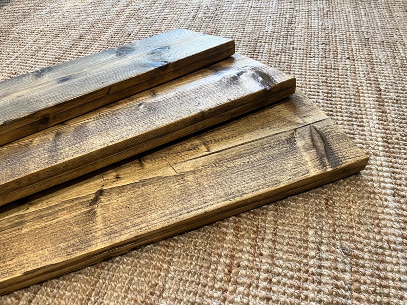 May include: Three rectangular wooden boards with a dark brown stained finish and visible wood grain patterns. The boards are stacked in a staggered formation, showcasing the natural texture, knots, and smooth edges of the finished timber planks.